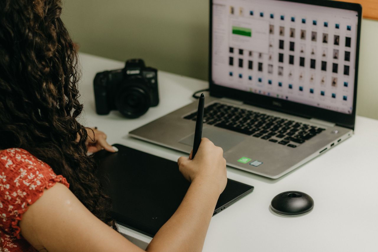 Woman using pen tablet with laptop and camera in creative workspace.